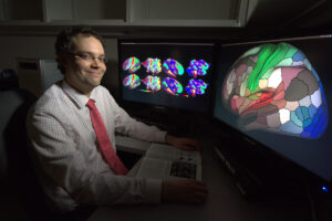 Matthew Glasser sits in front of two computer monitors displaying human brain cortical areas