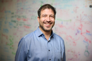 Dr. Adam Kepecs is a white man with short dark hair wearing a blue shirt in front of a white board with its contents blurred.