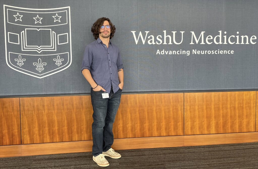 Leandro Fosque is a man wearing blue jeans and a purple button up shirt and glasses. He stands with hands in pocket in front of a sign that says WashU Medicine: Advancing neuroscience.