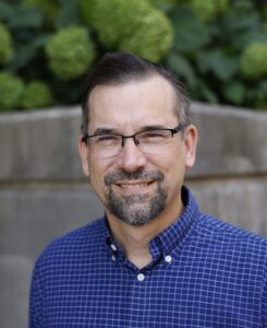 Steven Mennerick is a man with short gray and black hair and a black and gray beard. He wears glasses and a blue shirt.