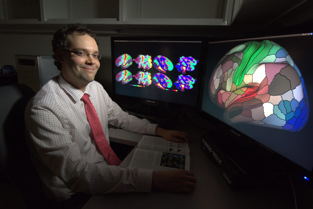 Matthew Glasser sits in front of two computer monitors displaying human brain cortical areas