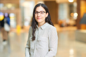 Kristen Prufrock is a woman with dark, longer hair and wearing glasses and a polka dot (dark dots on white) shirt.